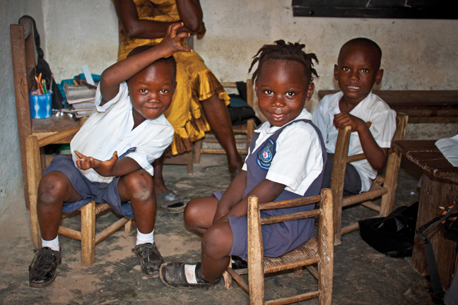 Three young children sit on wooden chairs in a classroom at The Salvation Army's Albert Orsborn Primary and Elementary School in Kakata, Liberia
