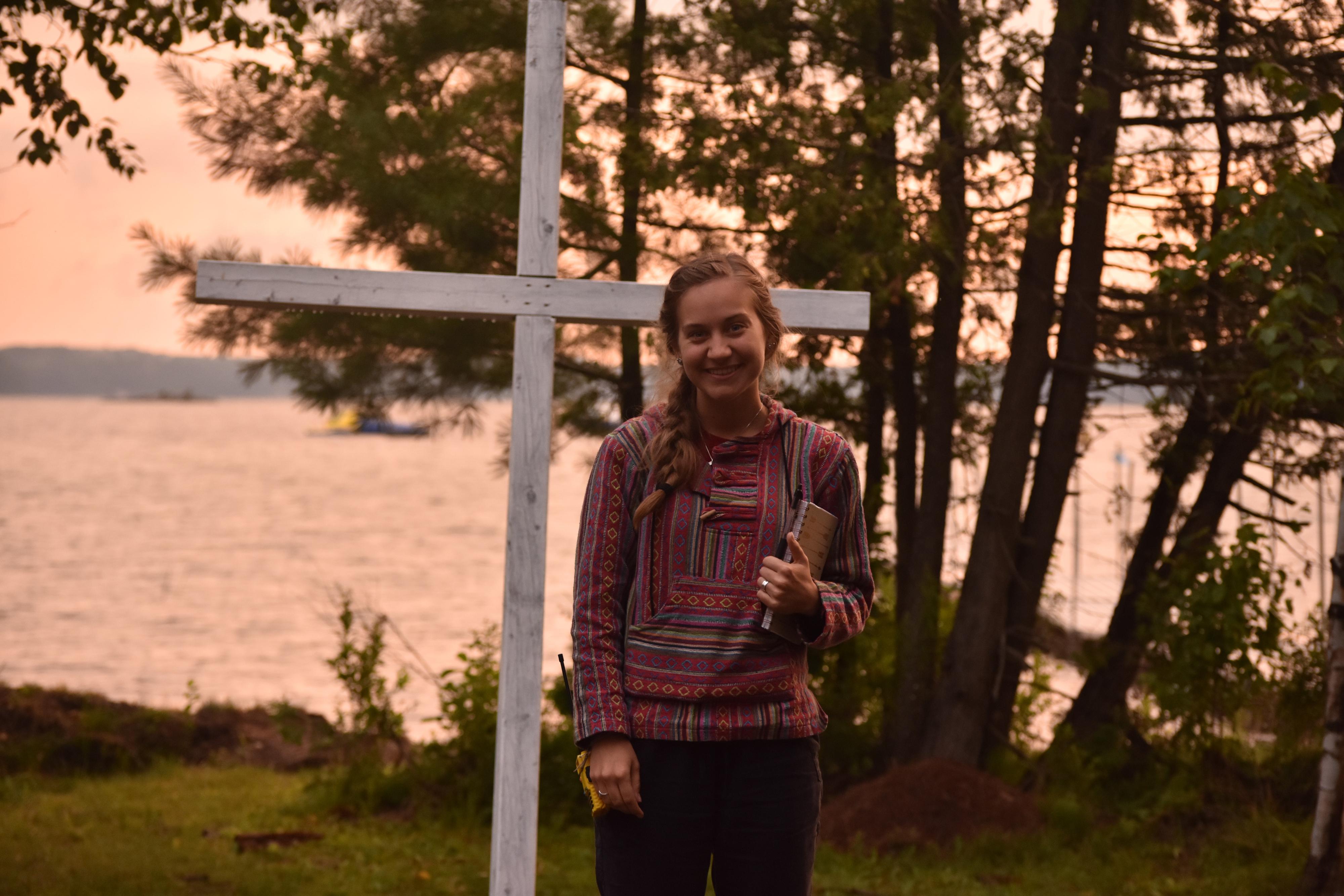 Misha Pavey stands in front of a white cross, holding a notebook, with lake in background