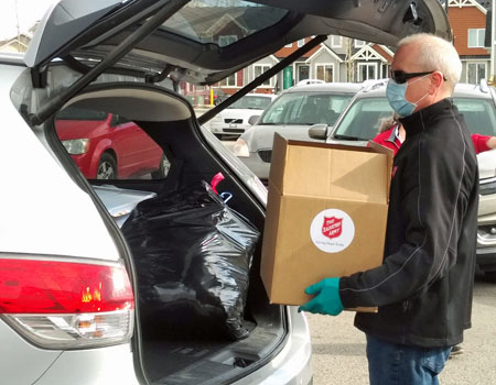 Salvation Army staff wearing a blue mask places a food hamper into the back of a vehicle