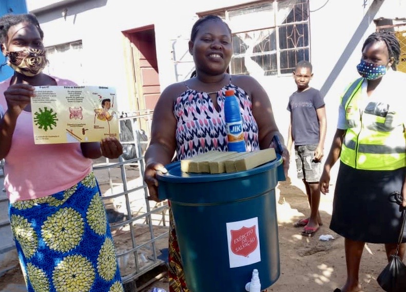 Three Salvation Army team members, one hold bucket of water, one a educational flyers about COVID-19, one stands with a yellow vest and mask. A child is in the background.