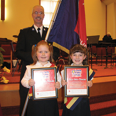 Alexandria (right) smiles on the day of her junior soldier enrolment alongside her cousin, Sarah Ball, and flag-bearer Kevin Thompson