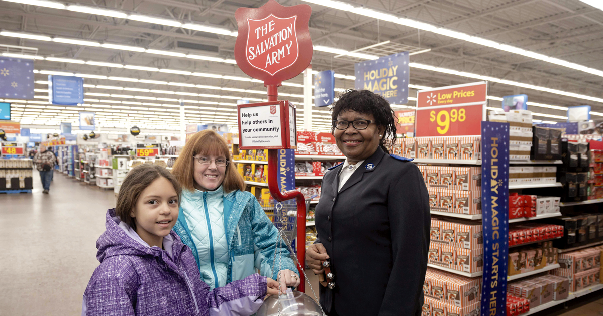 A Salvation Army soldier stands at a red kettle as smiling people donate