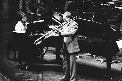 A man plays piano while another plays trombone