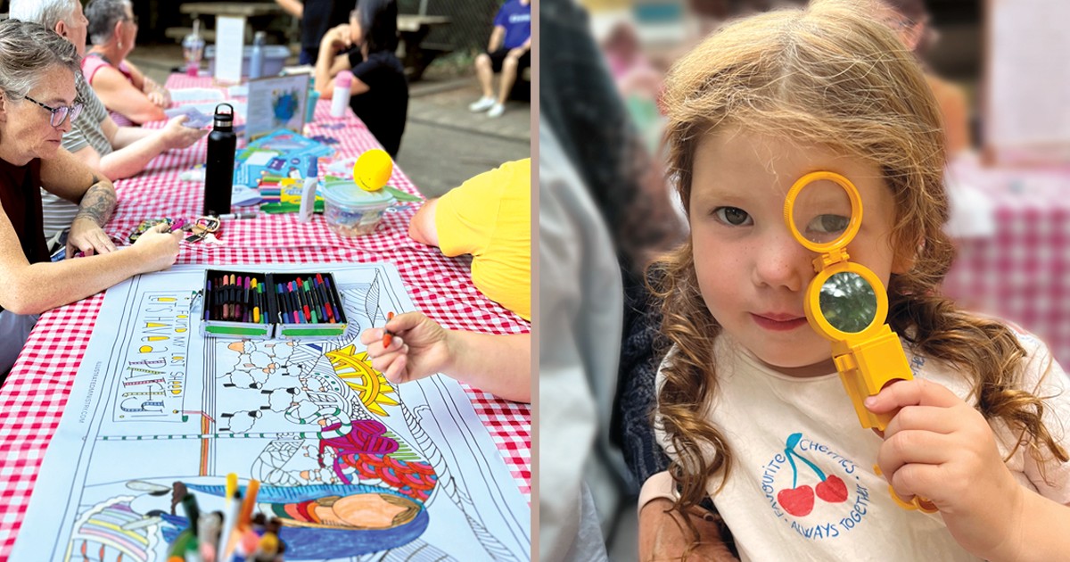 People colour a poster at a picnic table outside; a young girl holds a magnifying class up to her eye