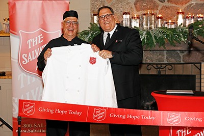 Two men standing and smiling, holding a chef's coat with the Salvation Army logo.