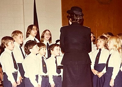 A group of children sing while a woman conducts